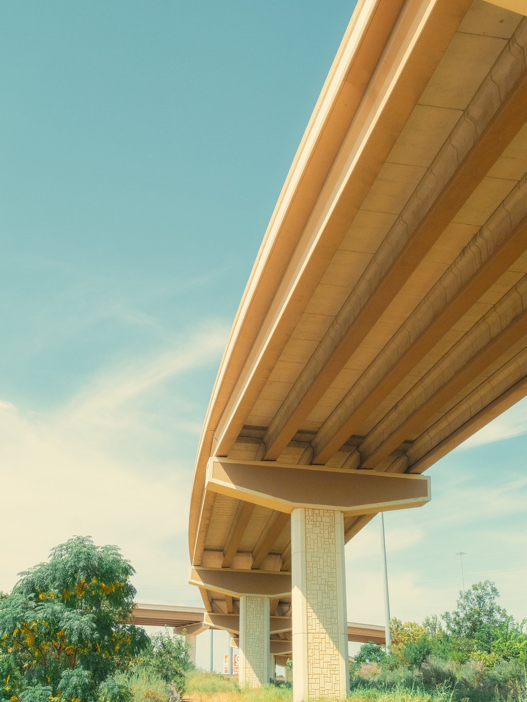 Low-angle view of a sunlit beige highway overpass with patterned concrete columns against a clear blue sky