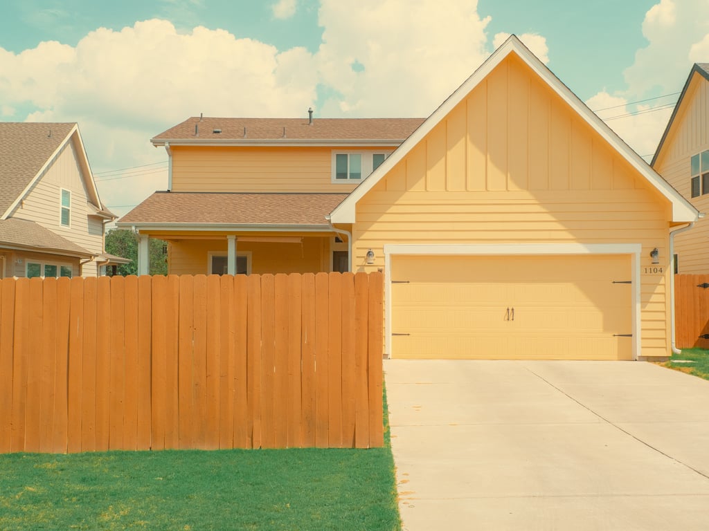 Sunny suburban view of a pale yellow house with an attached garage behind a tall wooden fence under a blue sky