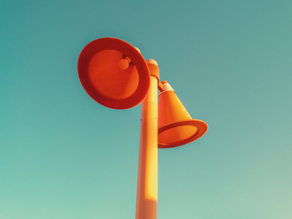 Three large orange megaphone-shaped horns mounted on a tall pole against a clear turquoise sky