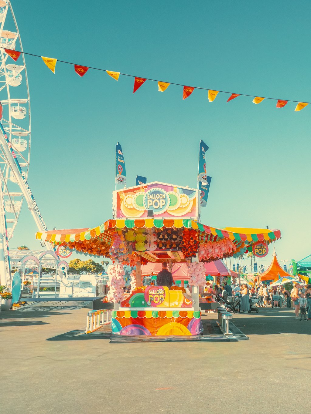 Bright carnival scene with a colorful Balloon Pop game stall, hanging stuffed prizes, and pennant flags under the sky