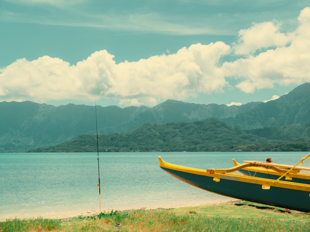 Yellow outrigger canoe on a grassy shore with turquoise sea stretching to dark tree-covered mountains beneath white clouds