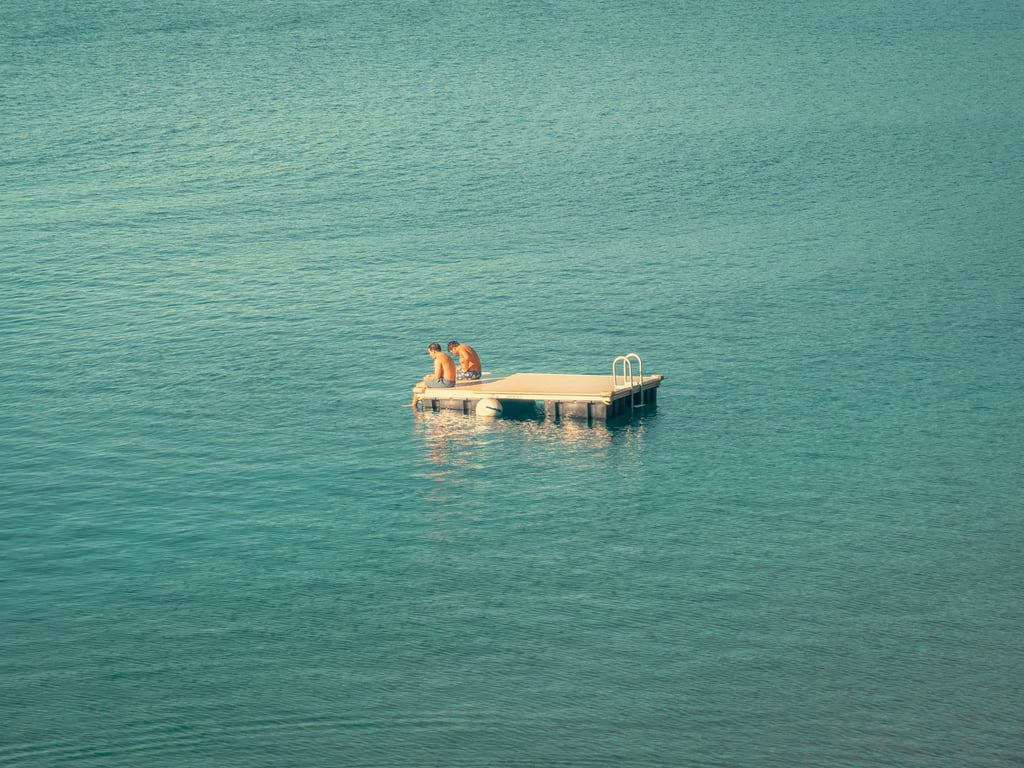 Two shirtless figures sitting on a small floating platform in a vast calm teal sea with a metal ladder and white buoy