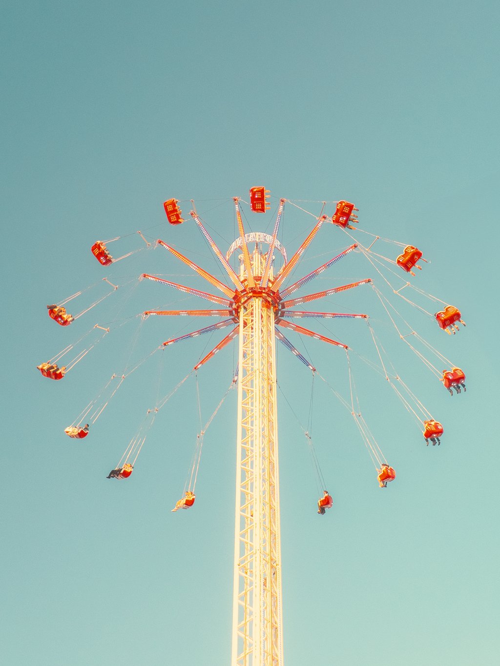 Tall vertical swing ride with a yellow lattice tower and red suspended seats spinning outward against a turquoise sky