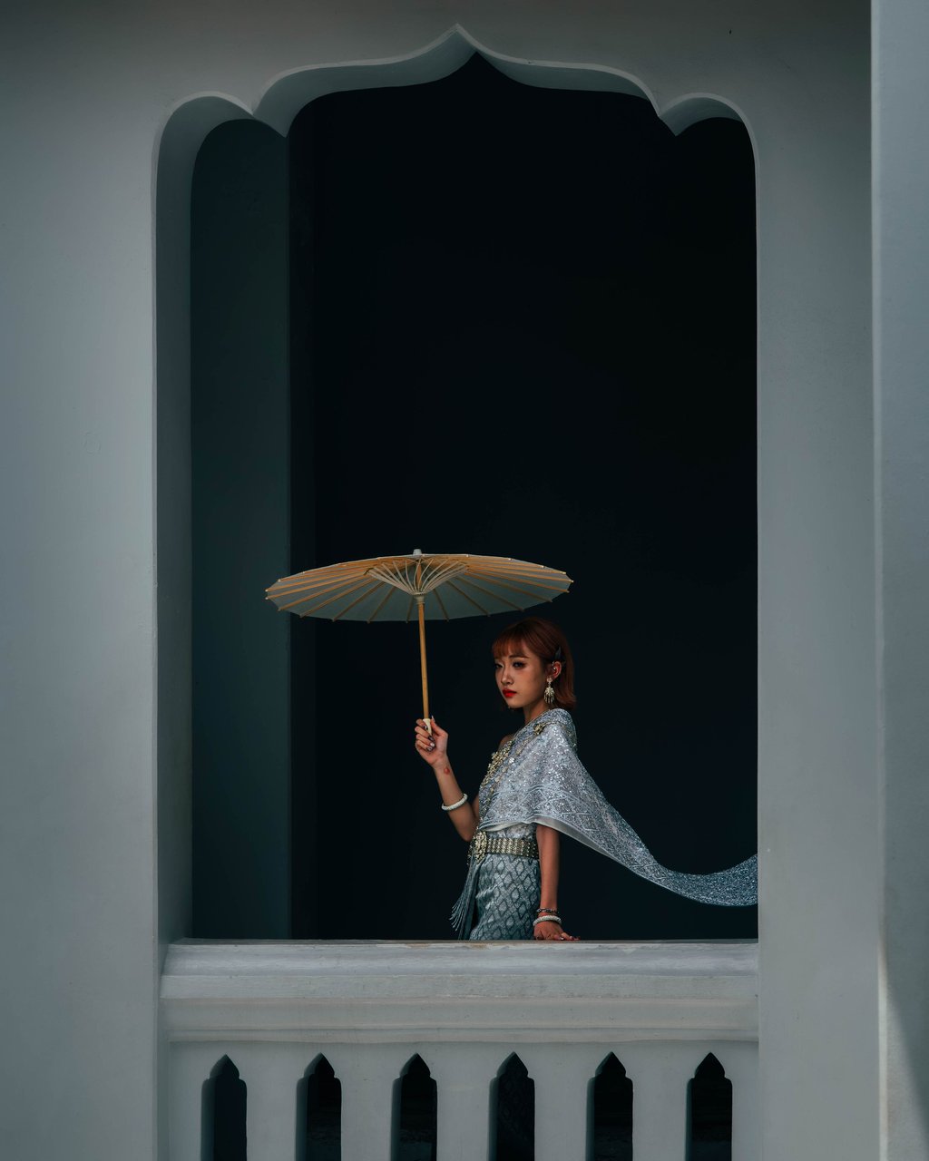 Woman in ornate patterned clothing holding a pale paper parasol in a dark arched window