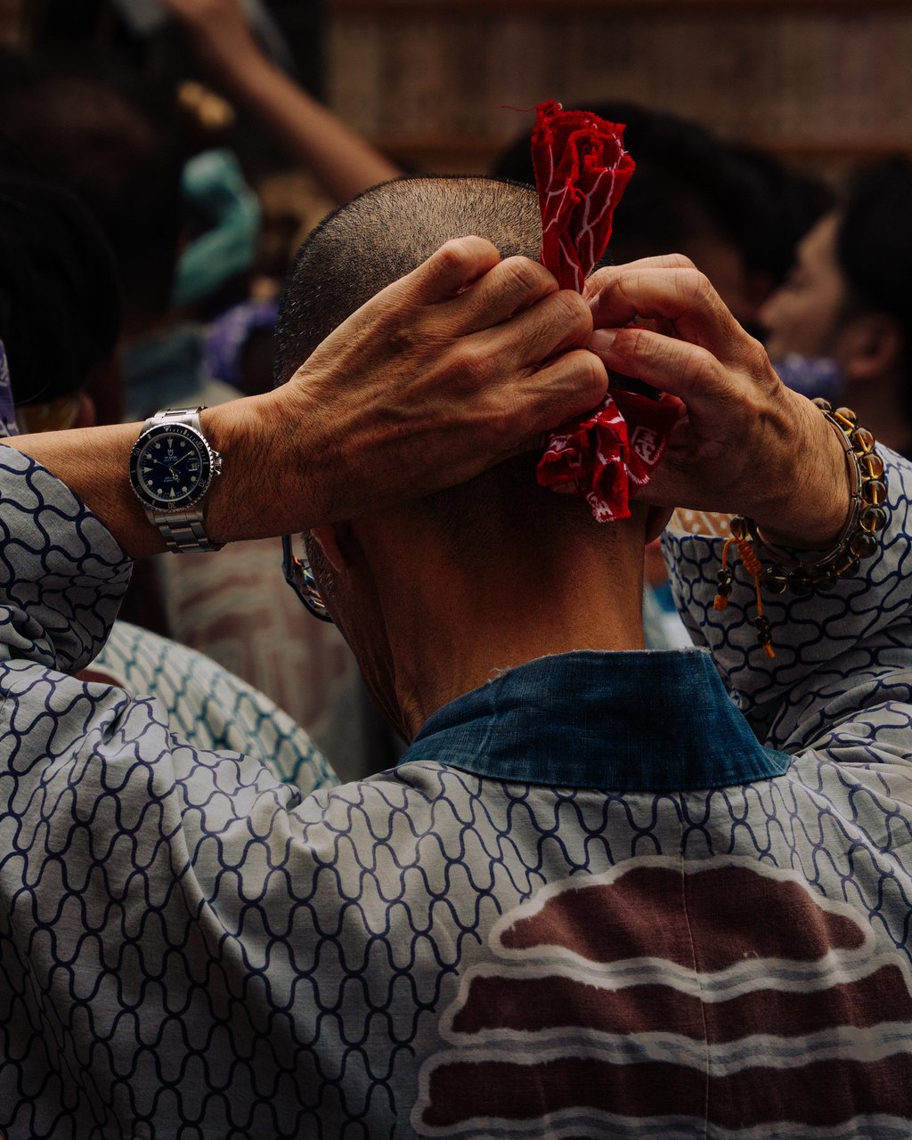 Close-up rear view of a shaved head as hands tie a red cloth at the nape with beaded bracelets