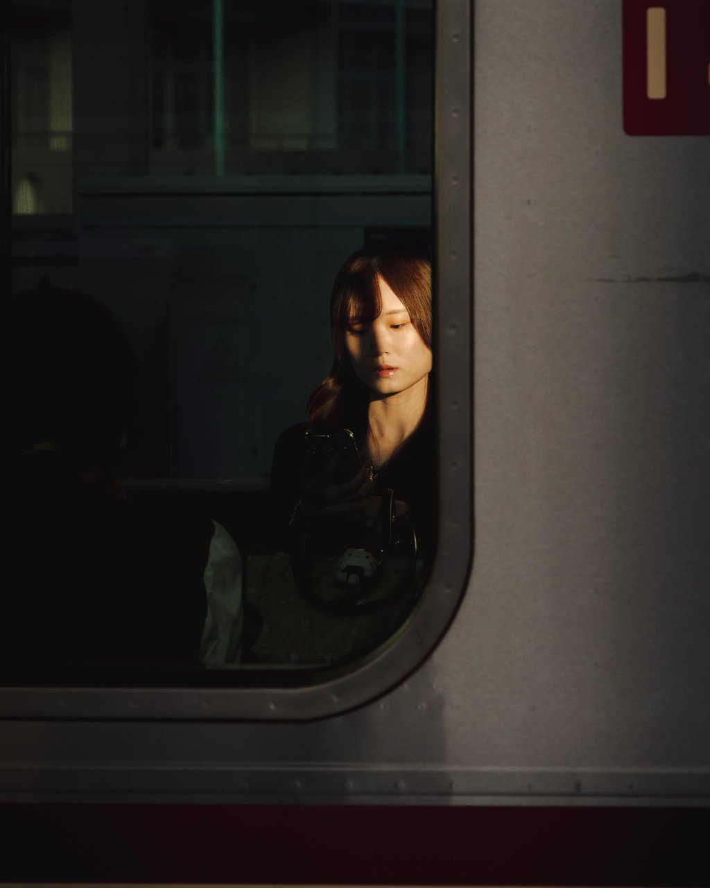 Moody view through a train window of a young woman half-lit by warm sunlight looking at her phone
