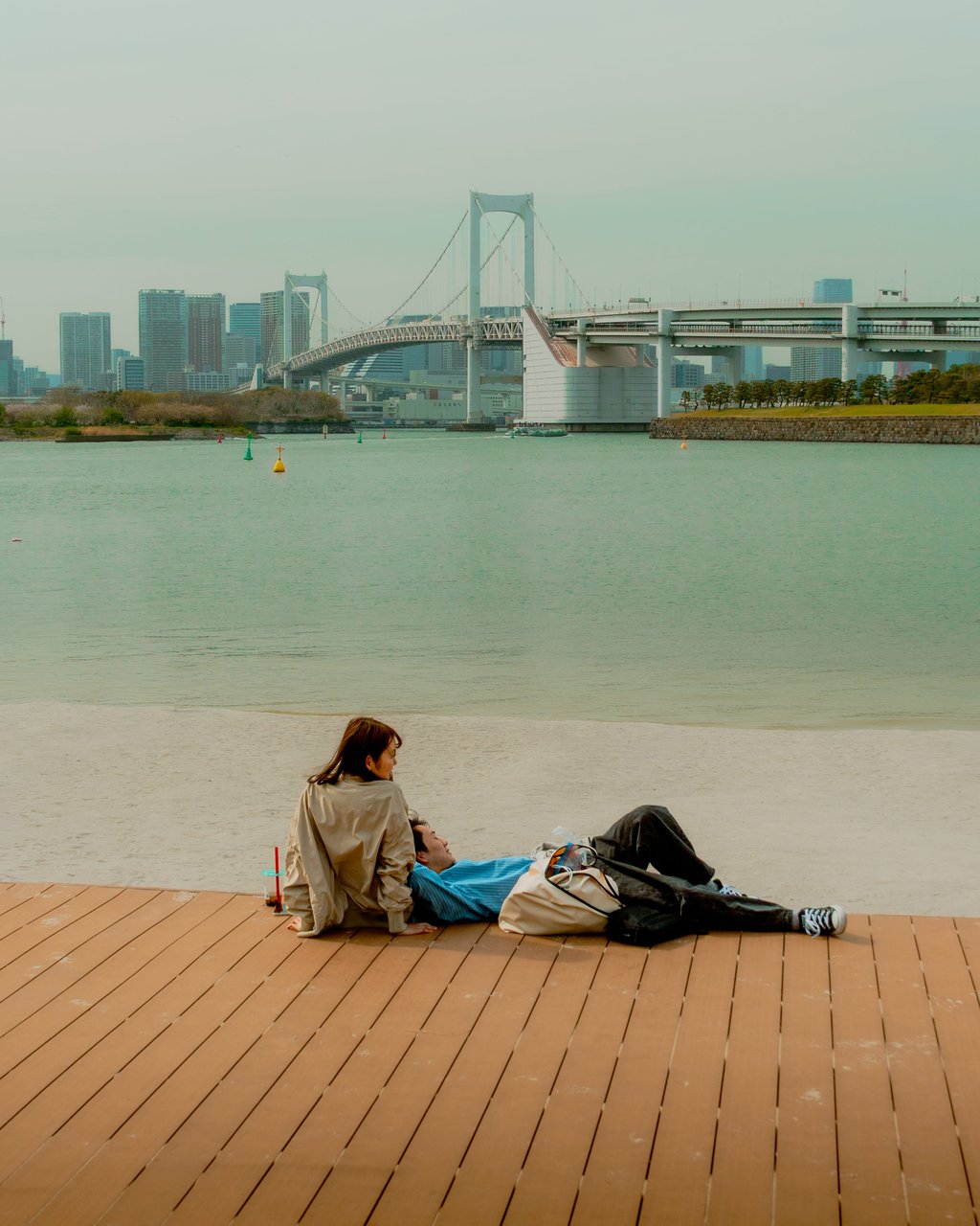 Two people relaxing on a wooden boardwalk at a calm waterfront with a suspension bridge in the background