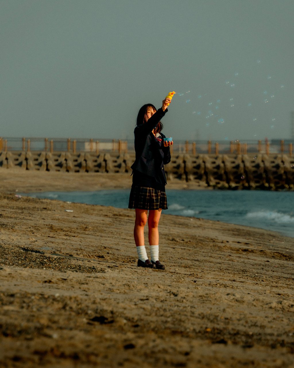 Person in a school uniform on a sandy beach blowing bubbles from a yellow duck-shaped bubble blower
