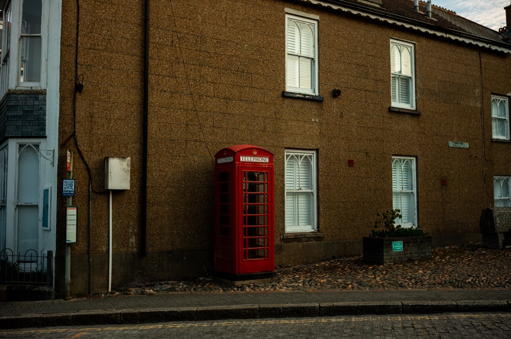 Red British telephone box against a pebble-dash building with shuttered white-framed windows on a cobbled pavement