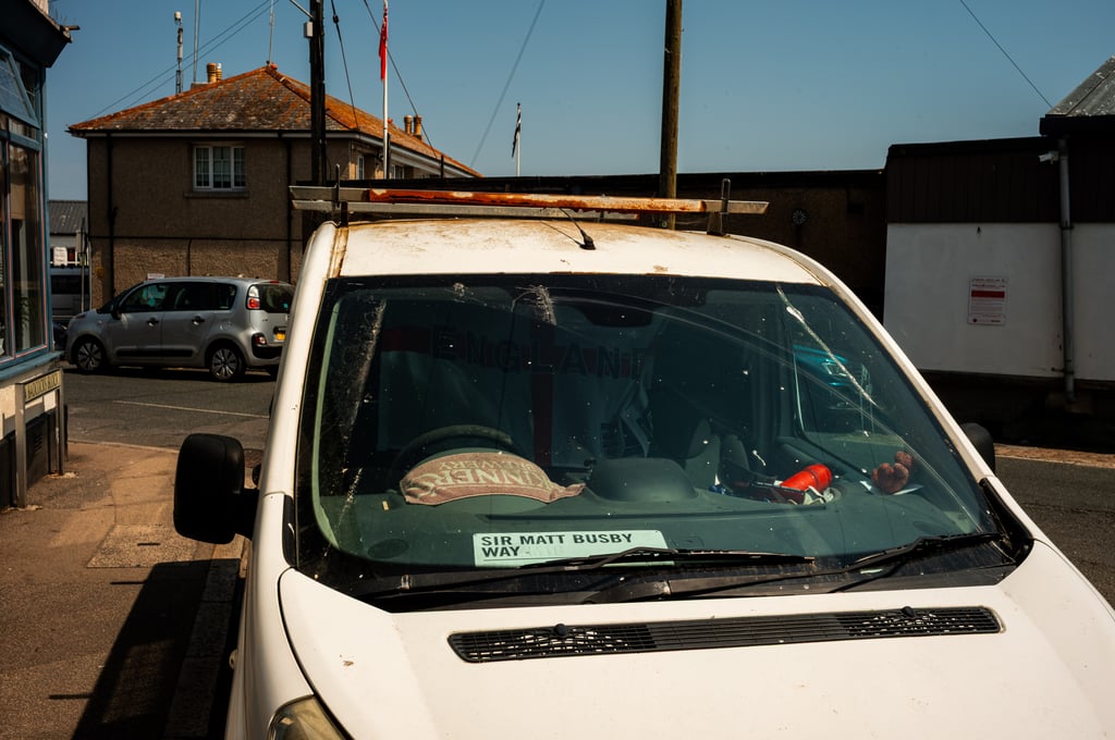 Front of a white van with a cracked windshield and rusted roof rack, a dashboard sign reading Sir Matt Busby Way