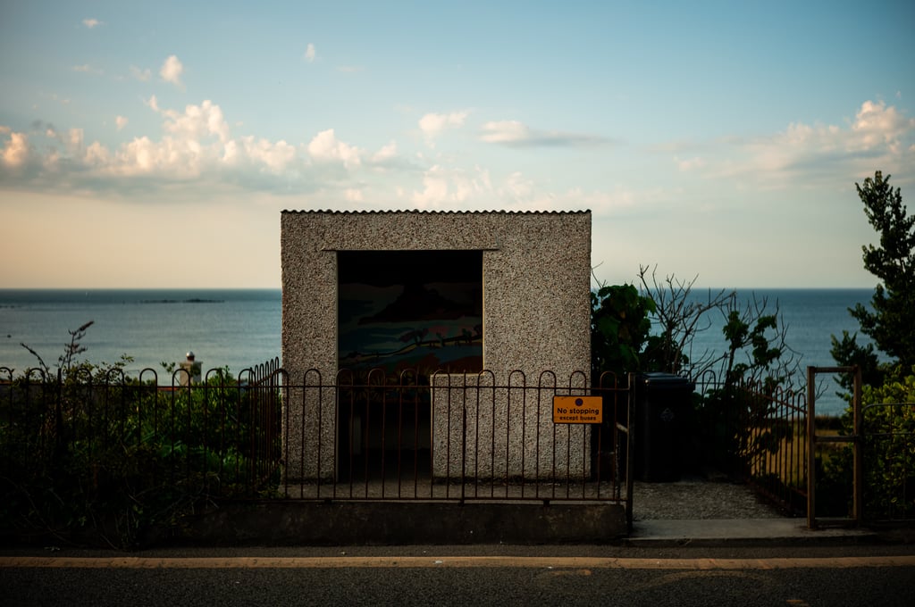 Small pebble-dash shelter with a gated iron fence overlooking the sea, a yellow sign reading No stopping except buses