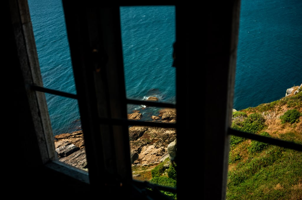 View through a dark window frame onto a turquoise sea and rocky shoreline with a grassy cliff slope