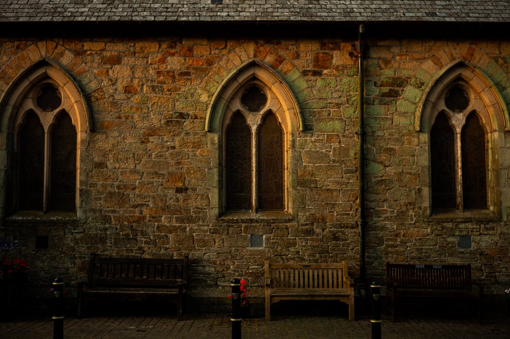 Weathered stone church facade with three pointed Gothic arched windows lit by warm sunlight and benches beneath