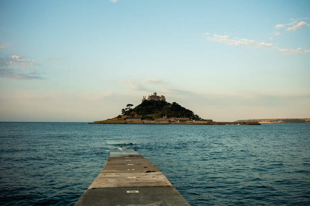 A concrete pier stretching across calm blue water toward a small tree-covered island topped by a stone castle