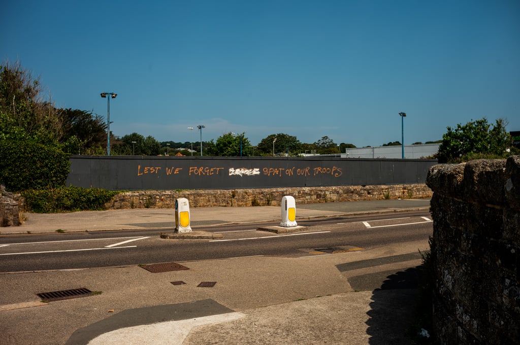 Street scene with a long grey barrier wall bearing orange graffiti reading Lest We Forget, with white traffic bollards