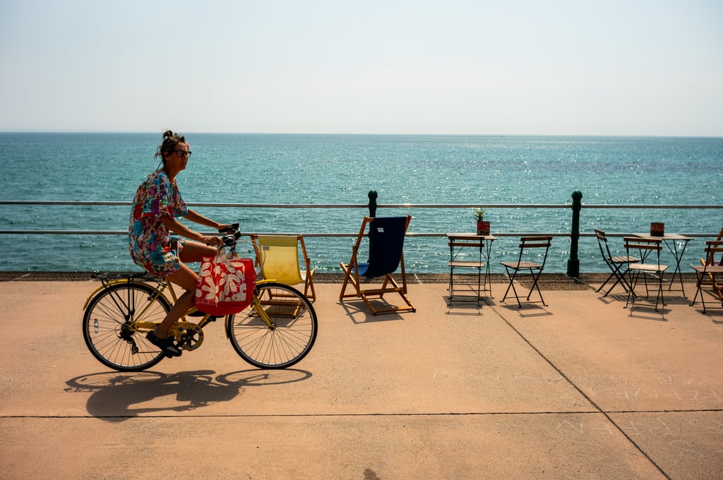 Person in a floral dress riding a yellow bicycle along a sunny seaside promenade lined with empty deck chairs