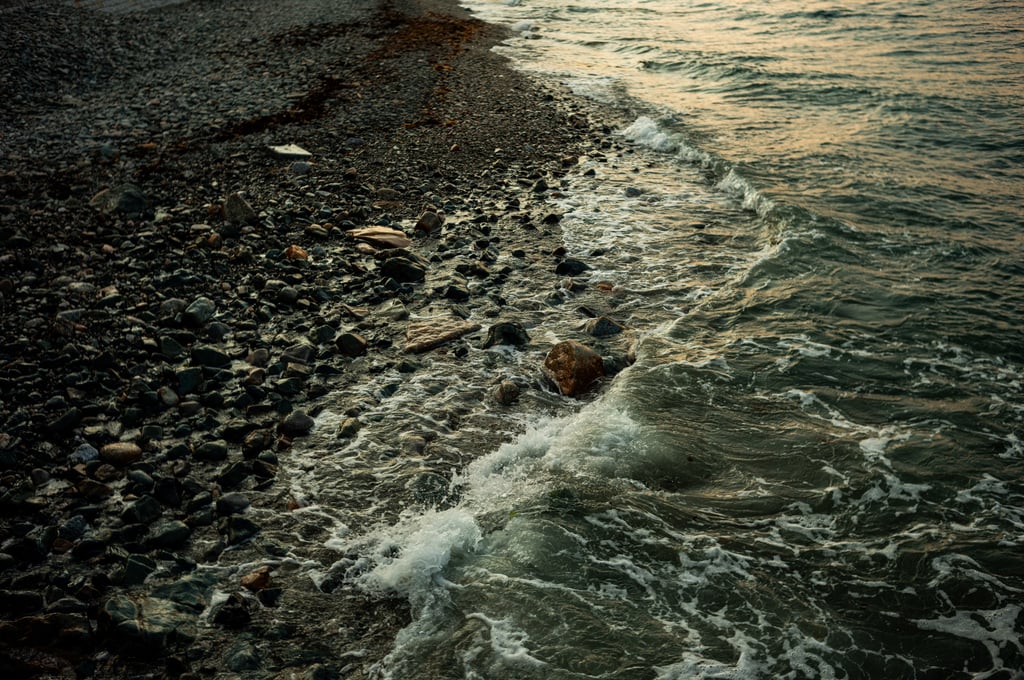 Close view of a rocky shoreline where small waves lap over wet pebbles, producing white foam on greenish sea