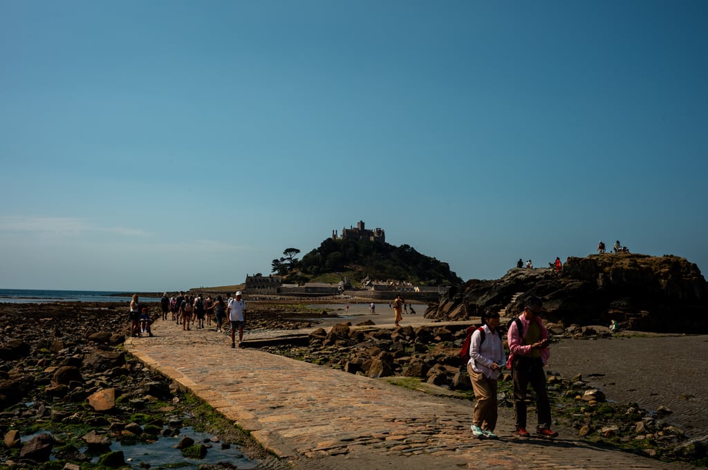 Crowds walking along a stone causeway across a rocky tidal shore toward a tree-topped island crowned by a castle