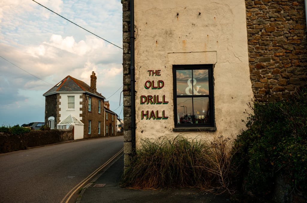 Weathered stone building painted The Old Drill Hall beside a window, on a quiet road with neighboring stone houses