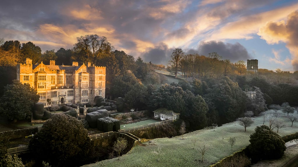 Aerial view of Fountains Hall surrounded by rolling hills beneath dramatic sunlit clouds.