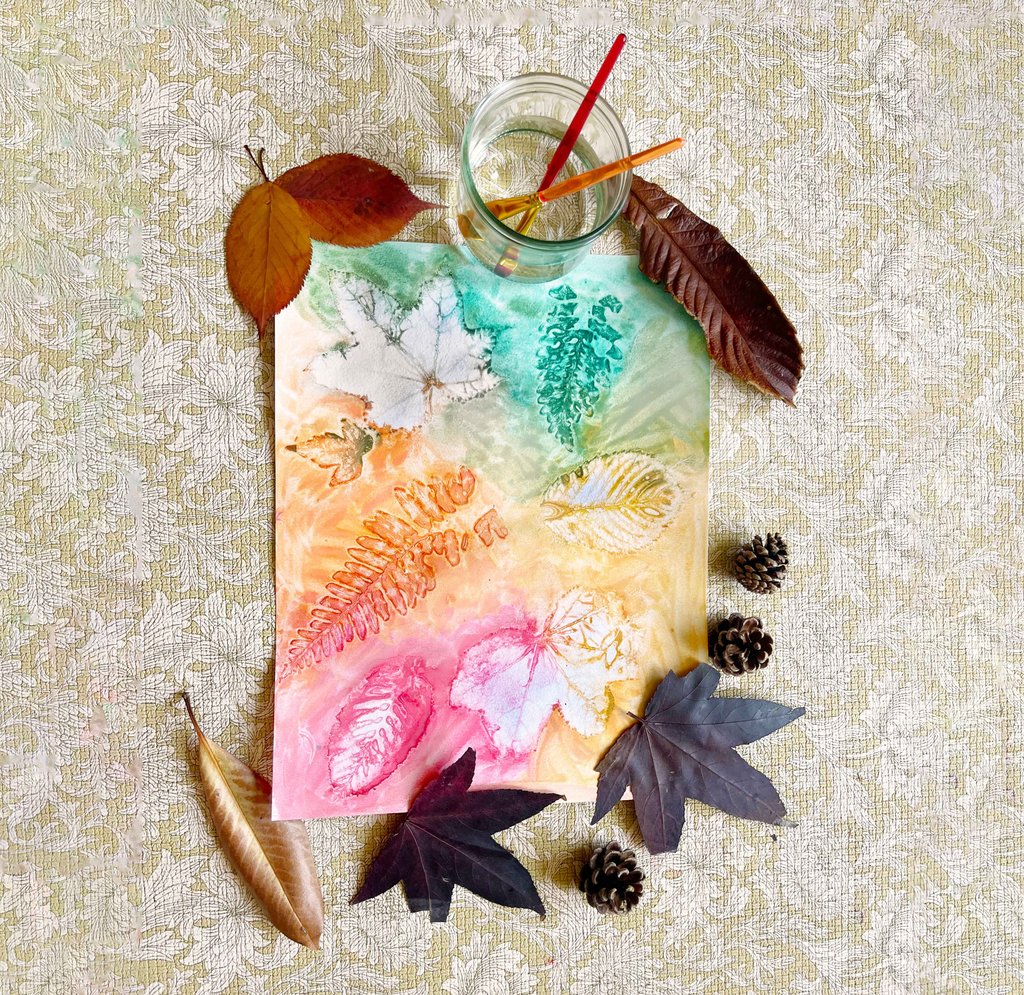 Watercolour painting of autumn leaves with pink, blue, and green splashes, surrounded by real leaves, a glass of water with paintbrushes, and art supplies.
