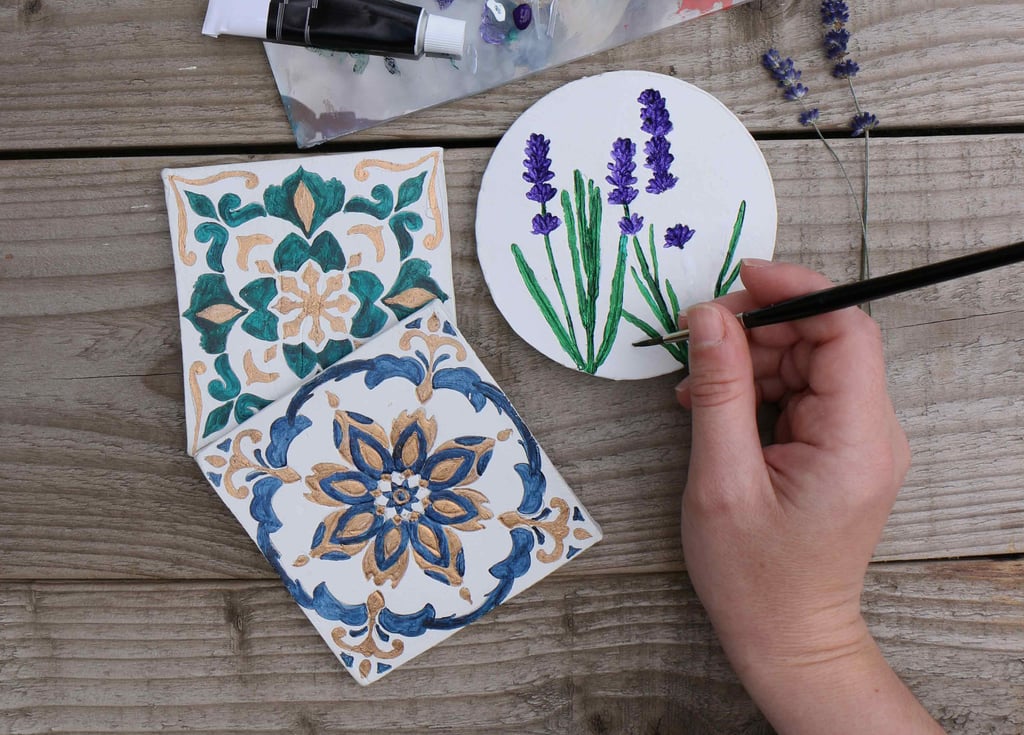 Person painting ceramic tile coasters at a wooden table with art supplies.